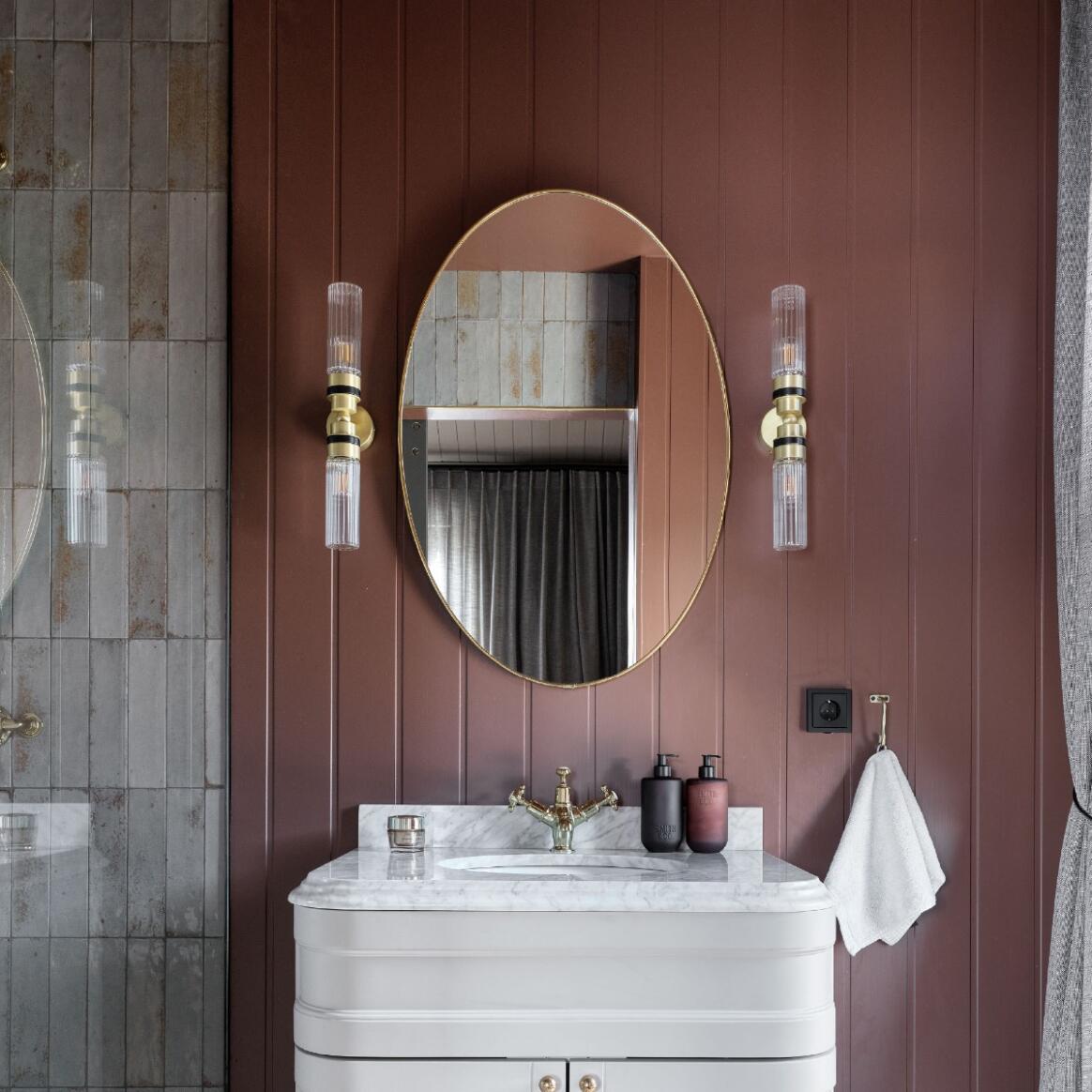 An oval wall mirror with a gold frame hangs above a white marble-top vanity in a bathroom with dark reddish-brown wood paneling. Two matching wall sconces flank the mirror, and a white towel hangs on the right.